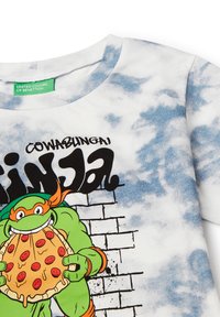 Bílý tričko s modrým tie-dye vzorem, které zobrazuje kreslenou želvu držící pizzu. Text „COWABUNGA NINJA!“ tučným černým písmem.