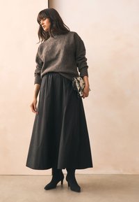 Brauner Strickpullover, schwarzer Maxi-Rock mit Kordelzugbund, schwarze Wildleder-Stiefeletten, gemusterte Clutch mit schwarz-weißen Akzenten.