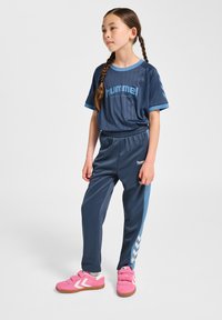 Dunkelblauer Trainingsanzug mit kurzärmligem Oberteil mit hellblauen Akzenten und Logo, kombiniert mit passenden Hosen. Helle pinke Sneaker vervollständigen das Outfit.