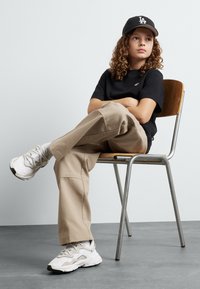 Maglietta nera, pantaloni beige a gamba larga e scarpe da ginnastica bianche. Capelli ricci, con un cappellino nero con logo LA, seduto su una sedia di metallo in un ambiente minimal.