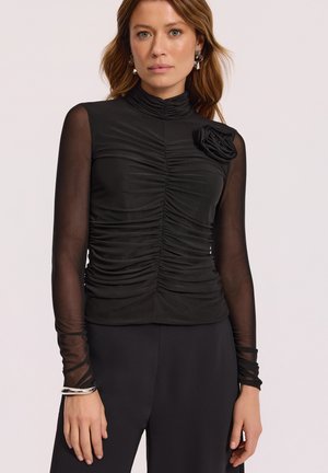 Kobieta ubrana w czarny marszczony top z przezroczystymi długimi rękawami, wysokim kołnierzem i ozdobą w postaci materiałowego kwiatu na lewym ramieniu, w połączeniu z czarnymi spodniami.