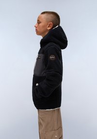 Veste en polaire noire avec une texture douce, une capuche et une fermeture éclair sur le devant. Présente des panneaux en blocs de couleurs et un logo rond sur la manche.