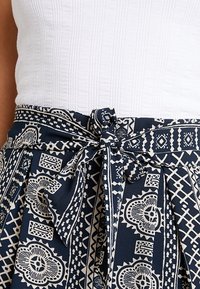 Marinblått mönstrat tyg med vita geometriska och blommig design, med ett knytbälte i midjan och en texturerad vit topp.