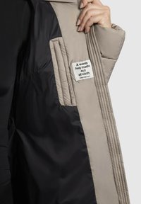 Bež zimska bunda z notranjostjo z odejano teksturo, črno podlogo in oznako, ki pravi "Topel objem iz blaga."