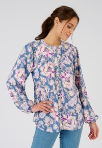 Camisa de manga larga con un patrón floral en azul, con acentos en púrpura y blanco, que presenta un escote redondo y cierre de botones en la parte delantera. Textura de tela suave.