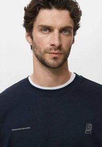 Man met blauwe ogen en bruin haar die een marineblauwe sweatshirt draagt met witte tekst "Crafting Culture Since 2002" en een "B" logo op een witte achtergrond.
