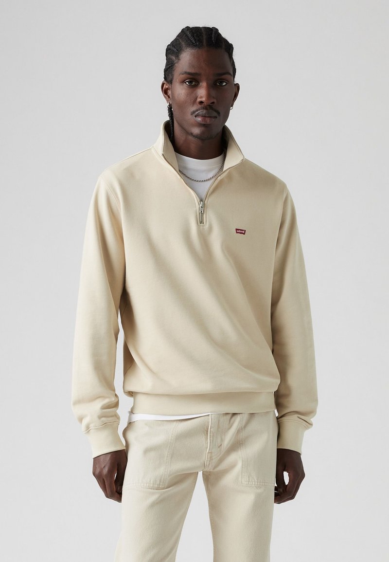 Mann mit geflochtenem Haar, trägt einen beigen Zip-Up-Sweatshirt, weißes Unterhemd, beige Hose und eine silberne Kette, steht vor einem schlichten Hintergrund.
