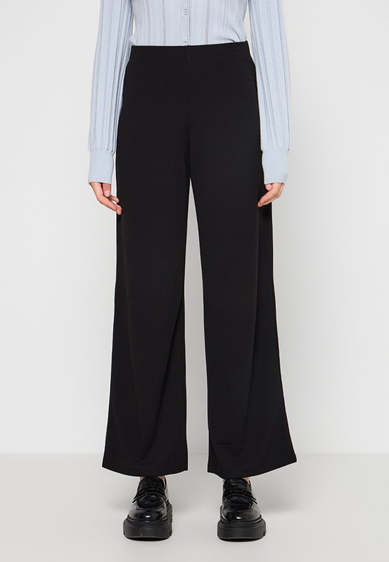 Pantaloni neri a gamba larga realizzati in tessuto morbido, con vita alta e dettagli minimi, abbinati a scarpe nere e a un top blu chiaro.
