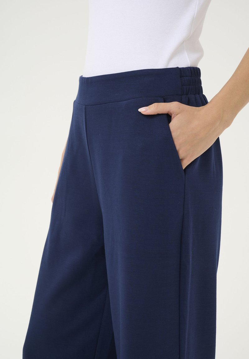 Marineblauwe broek van zacht materiaal, met een brede tailleband, zijzakken en een relaxte pasvorm. Gladde textuur met minimale naden.