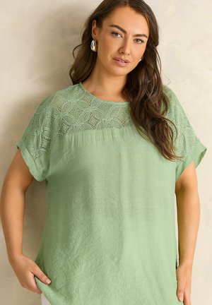 Kobieta z długimi brązowymi włosami, nosząca srebrne kolczyki koła i jasnooliwkową bluzkę z koronkowymi detalami na ramionach, stojąca na tle beżowej ściany.