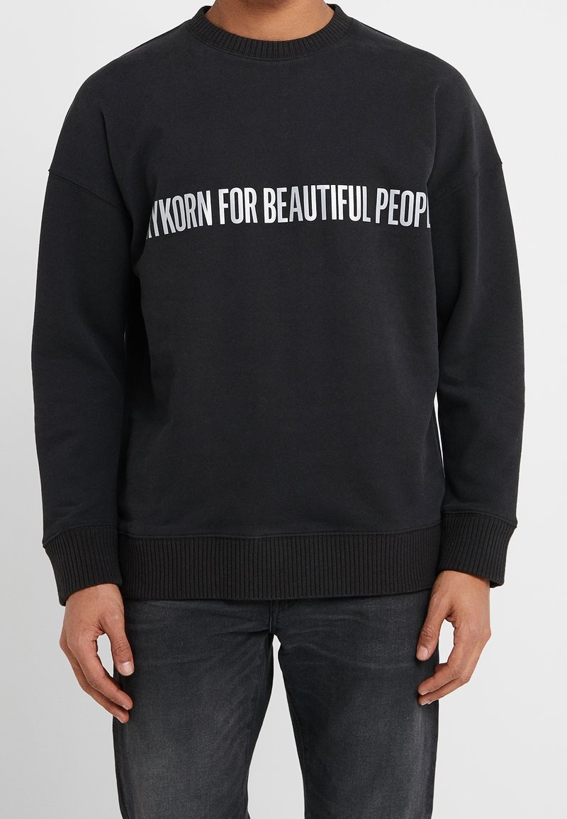 Personne portant un sweat-shirt noir avec un texte blanc partiellement visible, lisant "FOR BEAUTIFUL PEOPL,", associé à un jean foncé.
