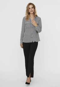 Donna in piedi che indossa una camicia a righe verticali bianca e nera con bottoni, pantaloni neri con spacchi frontali e tacchi neri su uno sfondo bianco.