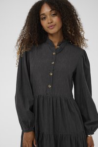 Juodos džinsinės suknelės su sluoksniuotu dizainu, raukšlėta iškirpte, ilgomis pūstomis rankovėmis ir mygtukų uždarymu priekyje. Minkšta tekstūra ir laisvas siluetas.