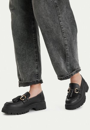 Person trägt schwarze klobige Loafer mit goldener Verzierung und Quasten, kombiniert mit hochgekrempelten, ausgewaschenen schwarzen Jeans vor hellem Hintergrund.