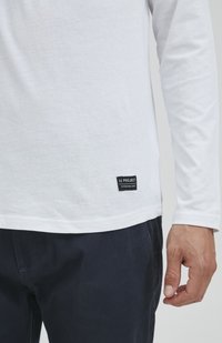 Witte T-shirt met lange mouwen van katoen. Heeft een zwart label met tekst aan de onderste zoom. Draagt met donkerblauwe broek.