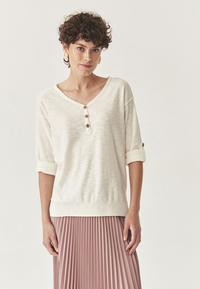 Helles beiges Pullover mit V-Ausschnitt, dreiviertel Ärmeln mit umgeschlagenen Abschlüssen und drei braunen Knöpfen. Strukturiertes Strickmuster und lockere Passform.