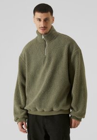 Fleece pulover u maslinasto zelenoj boji, s visokim ovratnikom, polu-razipom, rebrastim manžetama i porubom, s teksturiranom, prozirnom površinom.
