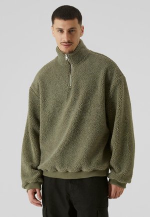 Fleece pullover i olivengrøn med høj krave, halvmatchende lynlås, ribbed cuffs og kant, samt en struktureret, gennemsigtig overflade.