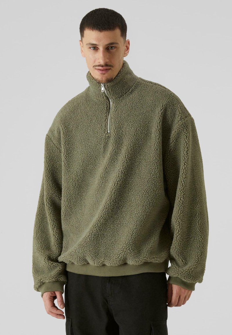 Fleece pulover u maslinasto zelenoj boji, s visokim ovratnikom, polu-razipom, rebrastim manžetama i porubom, s teksturiranom, prozirnom površinom.