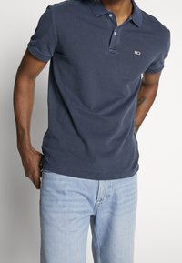 Polo blu navy in cotone, con chiusura a due bottoni, maniche corte e un piccolo logo sul petto. Vestibilità classica.