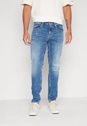 Homme portant un jean slim bleu clair légèrement usé, un t-shirt blanc et des baskets blanches, debout devant un fond uni.