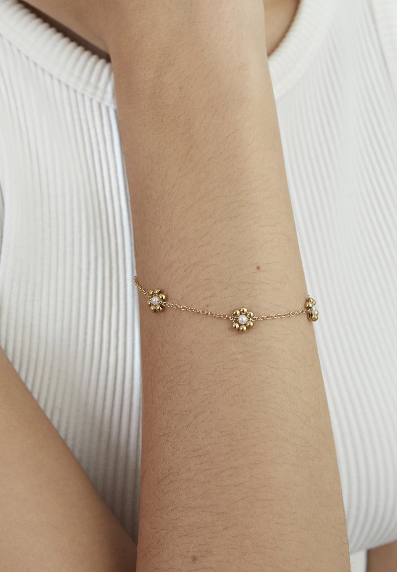 Bracciale a catena d'oro con tre ciondoli a forma di fiore con perle piccole, indossato su un avambraccio contro uno sfondo di top bianco a coste.