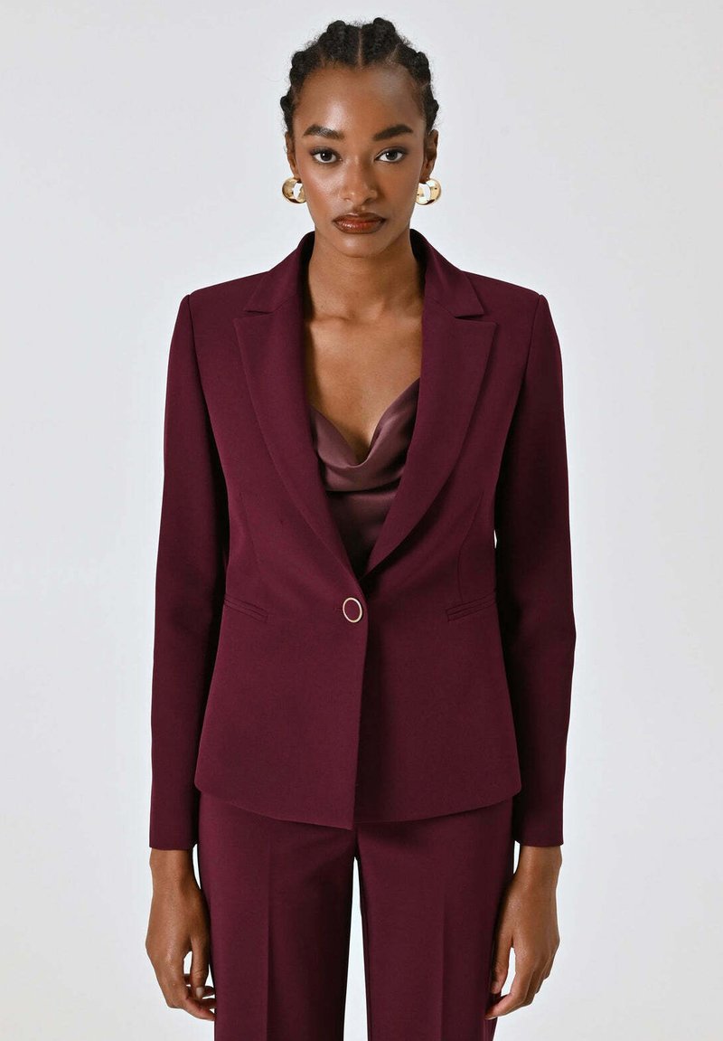 Blazer sartoriale color marrone scuro con un unico bottone dorato, rever a incavo e spalle imbottite. Tessuto liscio con vestibilità strutturata.