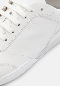 Valkoiset nahkakengät valkoisilla nauhoilla, näkyvillä ompelemilla ja kohokuvioitu "BOSS HUGO BOSS" -logo sivupaneelissa.