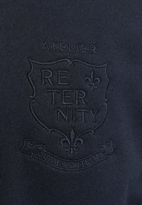 Tissu sombre avec blason en relief portant l'inscription "ATELIER RETERNITY" et des symboles décoratifs de fleurs de lys, ainsi qu'un texte en français sur un ruban en dessous.