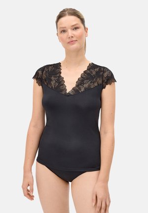 Mujer con un top ajustado negro con mangas cortas de encaje y escote en V, combinado con pantalones negros a juego, de pie frente a un fondo liso.