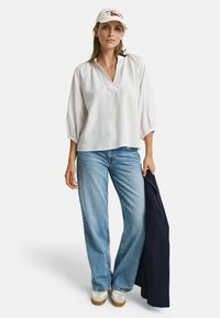Witte linnen blouse met V-hals, driekwart mouwen; gecombineerd met lichtblauwe spijkerbroek en witte sneakers. Een marineblauwe jas wordt vastgehouden.