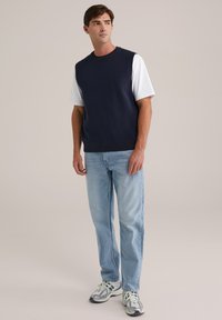 Mornarska trikotažna bezrukavna majica preko bijele T-majice, u kombinaciji s svijetloplavim ravnim trapericama i sivim sportskim cipelama s zelenim akcentima.