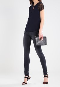 Haut bleu marine à manches courtes en dentelle, détail de cou en clé, associé  à un jean skinny noir et une pochette noire. Sandales à talons hauts noires.