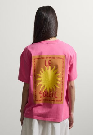 Person mit langen dunklen Haaren, die ein pinkfarbenes T-Shirt mit einer gelben Sonne und dem Text "LE SOLEIL" auf einem orangefarbenen Quadrat auf dem Rücken trägt.