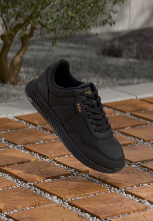 Basket noire BOSS avec semelle et lacets noirs flottant au-dessus d'une surface extérieure en bois carrelée et galets près des plantes.