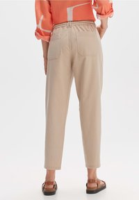 Pantalons beiges à jambe droite avec une taille élastique, deux poches arrière et une texture lisse. Portés avec des sandales marron sur un fond clair.