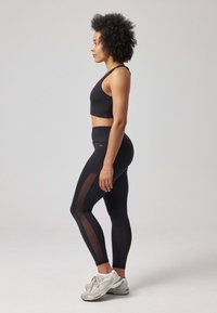 Soutien-gorge de sport noir et leggings avec panneaux latéraux transparents, dotés d'une taille haute et d'un tissu léger et extensible. Modèle portant des baskets neutres.