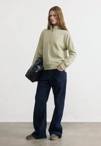 Γυναίκα που φοράει μπεζ φούτερ με φερμουάρ στο ένα τέταρτο, σκούρο τζιν με φαρδύ παντελόνι, γκρι αθλητικά παπούτσια, κρατώντας μαύρη τσάντα, στέκεται μπροστά σε απλό λευκό φόντο.