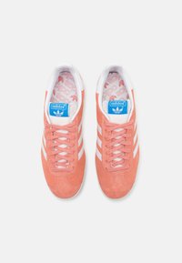 Sneaker in suede rosa con dettagli bianchi, caratterizzati da tre strisce laterali, punta arrotondata e logo Adidas blu sulla linguetta.