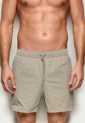 Męski tors bez koszulki, ubrany w beżowe szorty kąpielowe ze ściągaczem, stojący na jednolitym tle.