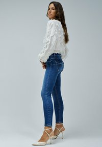 Blusa blanca texturizada con detalles de volantes, combinada con jeans azul ajustados y zapatos de tacón beige. Fondo gris sencillo.