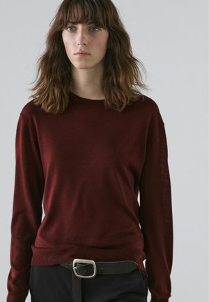 Pull en tricot bordeaux à manches longues et col rond. Associé à une ceinture foncée avec une boucle rectangulaire argentée.