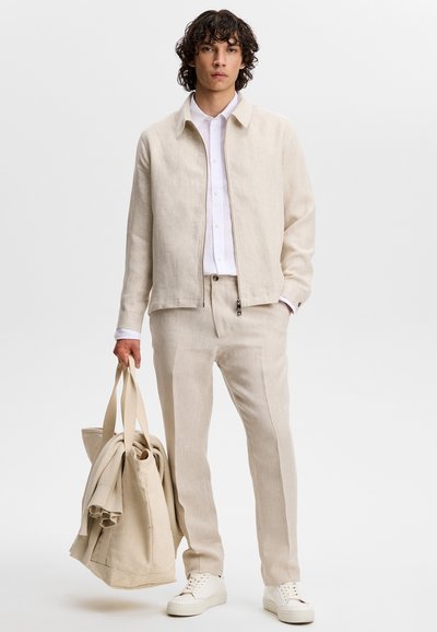 Άνδρας με μπεζ σακάκι και παντελόνι, λευκό πουκάμισο και αθλητικά παπούτσια, κρατώντας μια τσάντα tote σε αντίστοιχο μπεζ χρώμα και ένα διπλωμένο ένδυμα.