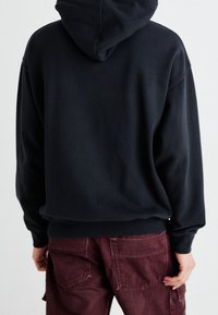 Sudadera negra con bolsillo canguro, corte oversize y capucha con cordón, combinada con pantalones cargo burdeos que tienen múltiples bolsillos.