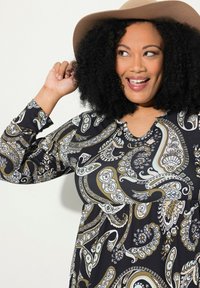 Musta mekko, jossa on beige, sininen ja valkoinen paisley-kuvio; siinä on pitkät hihat ja V-pääntie. Mallilla on päässään vaalea beige hattu.