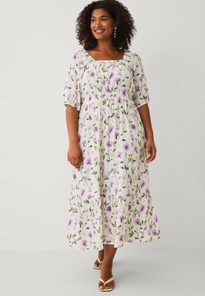 Smilende kvinde iført hvid midi-kjole med lilla blomsterprint, pufærmer, firkantet halsudskæring og beige sandaletter med åben tå, stående på ensfarvet baggrund.
