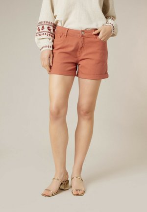 Femme portant un short rouille à ourlet roulé, un pull beige avec broderies rouges sur les manches, et des sandales à talons blocs dorés.