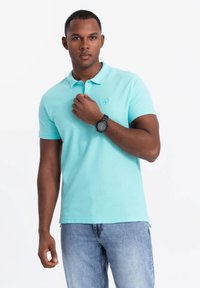 Světle modré polo tričko s malým vyšitým logem. Vydáno s vybledlými modrými džíny. Model má na levém zápěstí černé hodinky.