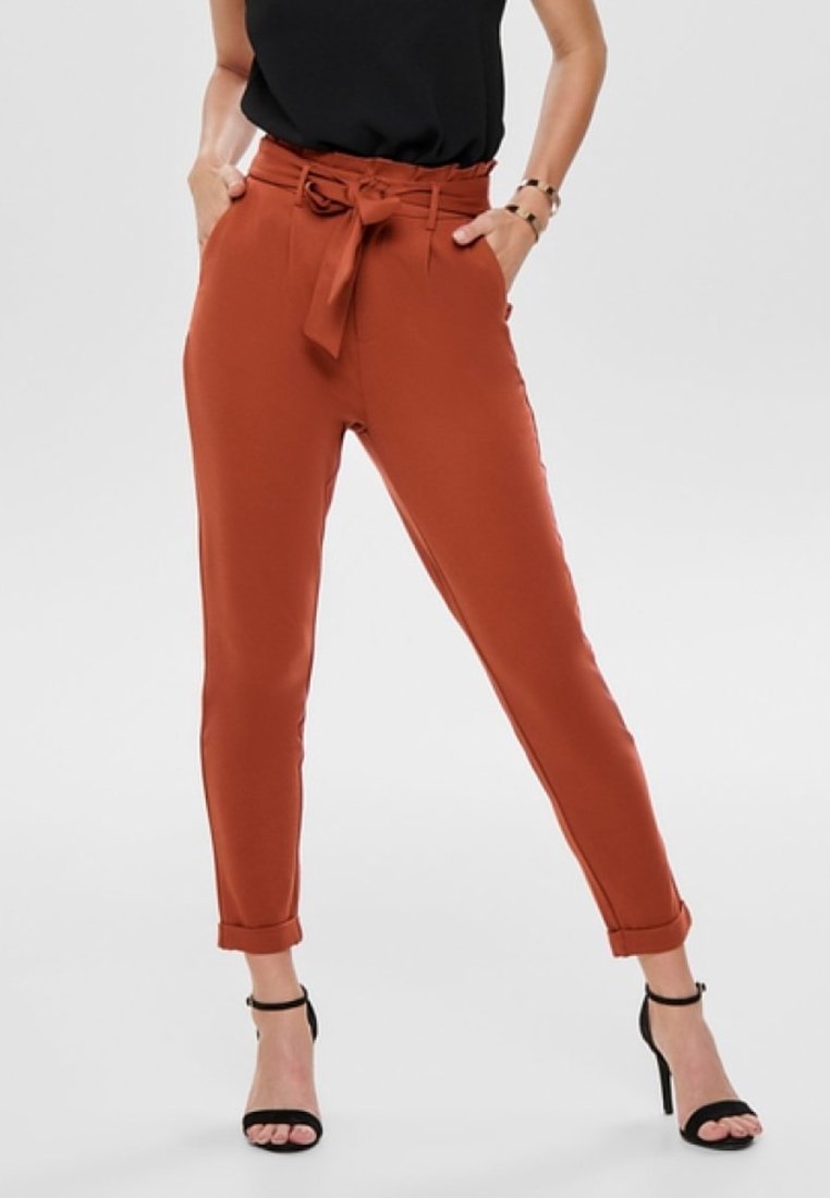 Femme portant un pantalon taille haute couleur rouille avec ceinture nouée, top noir sans manches, sandales noires à talons hauts, mains dans les poches, fond blanc.
