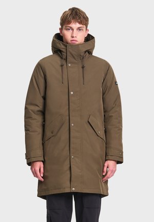 Jeune homme portant un long manteau d'hiver vert olive avec une capuche, des boutons sur le devant et des poches à rabat, debout devant un fond clair uni.